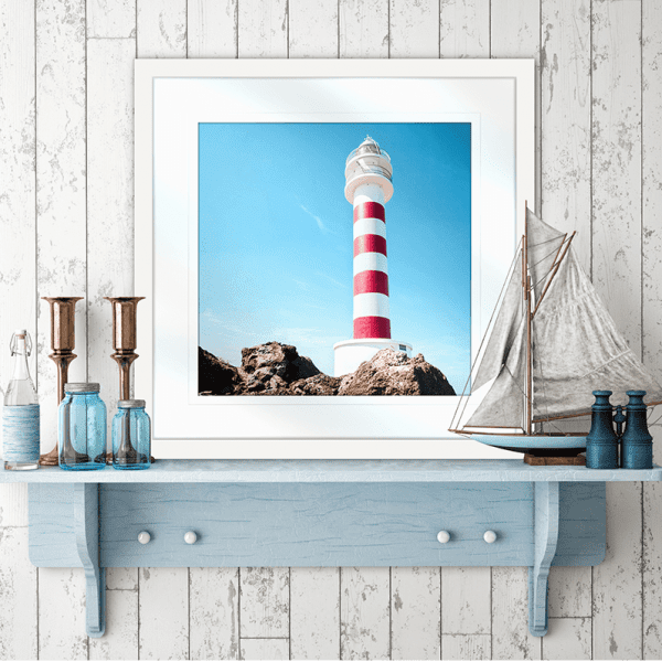Wall Mounted White framed Coastal Wall Art featuring a red and white lighthouse perched above the rocks, against a stunning blue sky backdrop