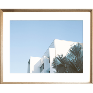 Gold framed Tropical Wall Art - The Weekender - Artist Collection, featuring striking a crisp white tropical hotel nestled amongst breezy palm trees, against a sky blue background.