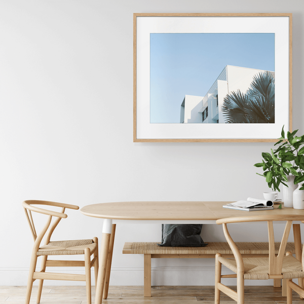 Wall mounted gold framed Tropical Wall Art - The Weekender - Artist Collection, featuring striking a crisp white tropical hotel nestled amongst breezy palm trees, against a sky blue background.