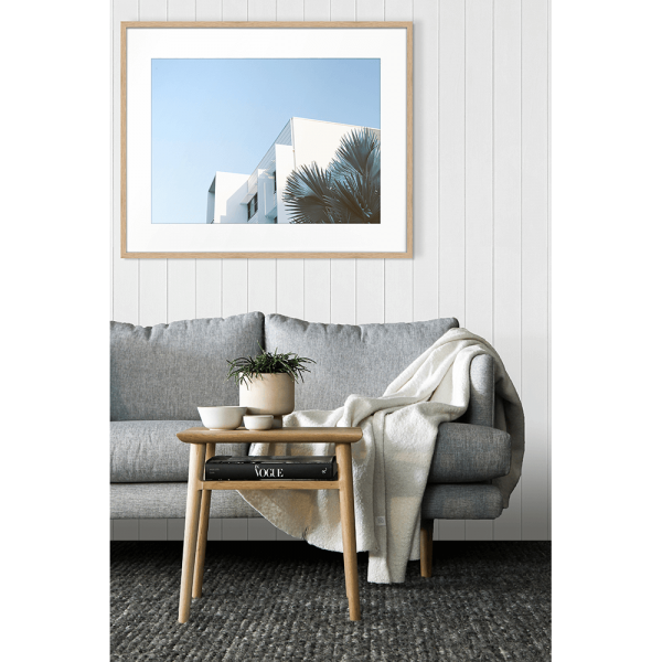 Gold framed in a lunge setting Tropical Wall Art - The Weekender - Artist Collection, featuring striking a crisp white tropical hotel nestled amongst breezy palm trees, against a sky blue background.
