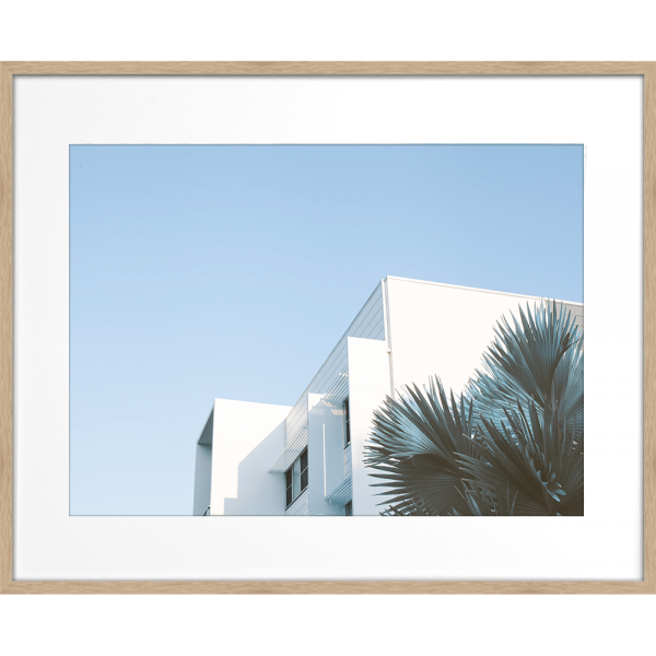 Wood framed Tropical Wall Art - The Weekender - Artist Collection, featuring striking a crisp white tropical hotel nestled amongst breezy palm trees, against a sky blue background.