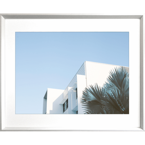 Silver framed Tropical Wall Art - The Weekender - Artist Collection, featuring striking a crisp white tropical hotel nestled amongst breezy palm trees, against a sky blue background.