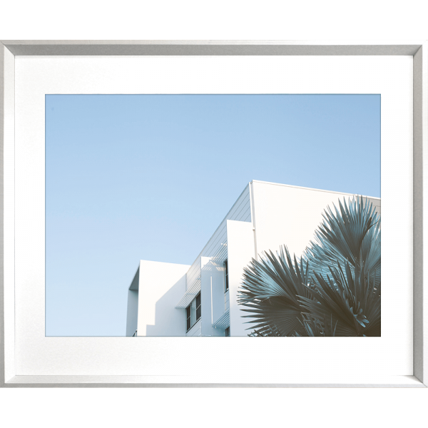 Silver framed Tropical Wall Art - The Weekender - Artist Collection, featuring striking a crisp white tropical hotel nestled amongst breezy palm trees, against a sky blue background.