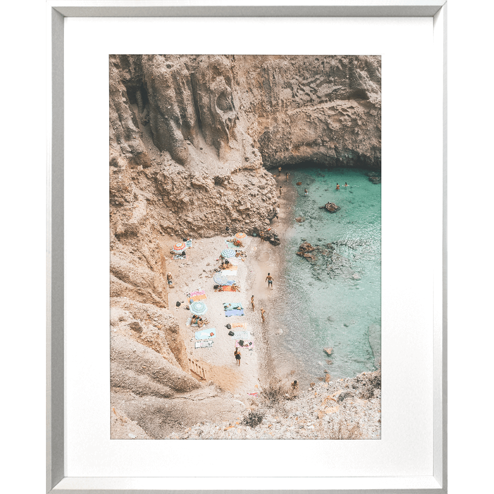 Silver framed Coastal Wall Art - Vacanza 01 – Studio Collection, Featuring a relaxing beach scene nestled within a cliff face. Clear aqua water with beach lovers sun baking in the sunshine.