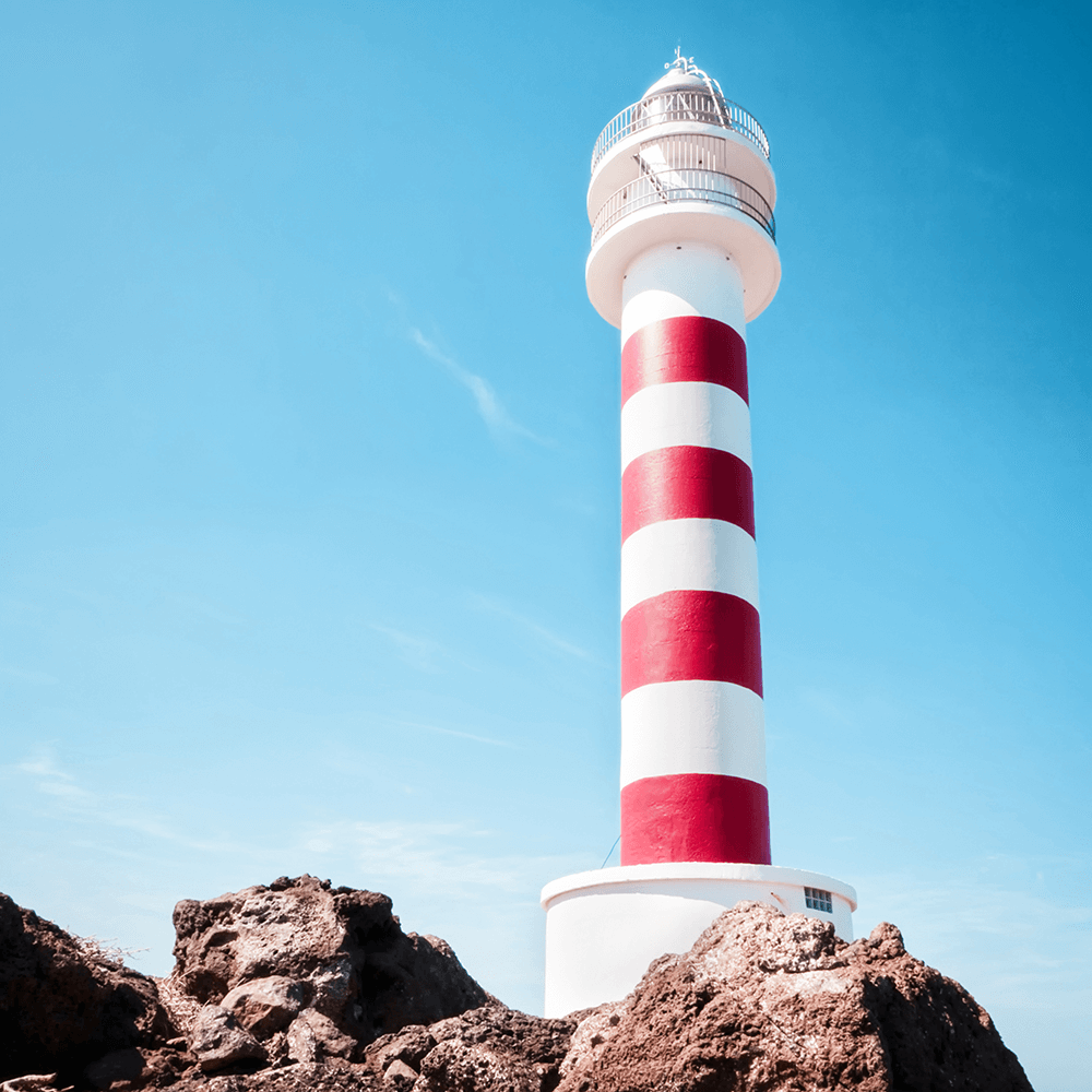 Unframed Coastal Wall Art featuring a red and white lighthouse perched above the rocks, against a stunning blue sky backdrop