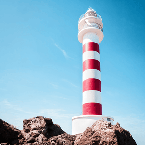 Unframed Coastal Wall Art featuring a red and white lighthouse perched above the rocks, against a stunning blue sky backdrop