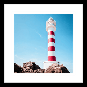 Black framed Coastal Wall Art featuring a red and white lighthouse perched above the rocks, against a stunning blue sky backdrop