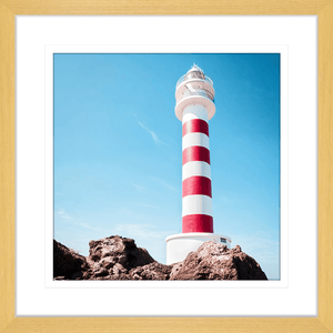 Gold framed Coastal Wall Art featuring a red and white lighthouse perched above the rocks, against a stunning blue sky backdrop