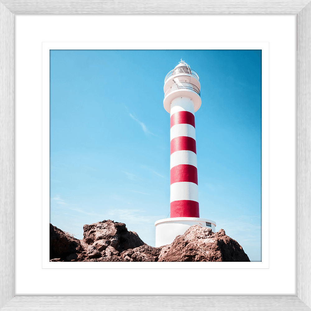 Silver framed Coastal Wall Art featuring a red and white lighthouse perched above the rocks, against a stunning blue sky backdrop