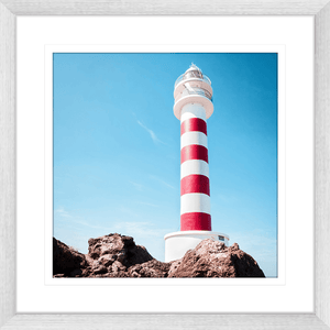 Silver framed Coastal Wall Art featuring a red and white lighthouse perched above the rocks, against a stunning blue sky backdrop