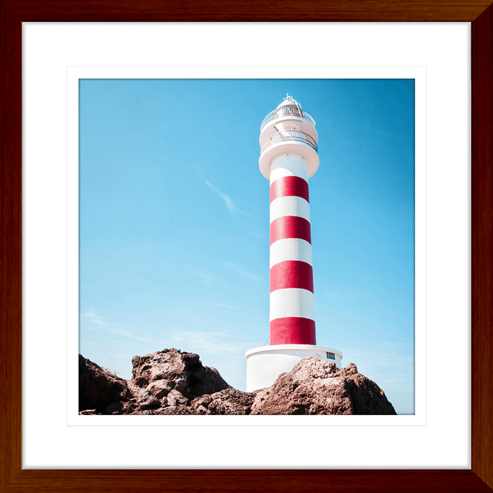 Wood framed Coastal Wall Art featuring a red and white lighthouse perched above the rocks, against a stunning blue sky backdrop