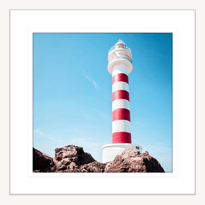 White framed Coastal Wall Art featuring a red and white lighthouse perched above the rocks, against a stunning blue sky backdrop