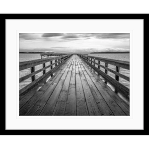 Black and white wall art features a long pier cascading into the deep ocean, with an array of white clouds overhead in a black frame