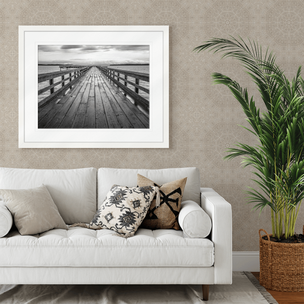 Black and white wall art features a long pier cascading into the deep ocean, with an array of white clouds overhead, wall mounted in a white frame