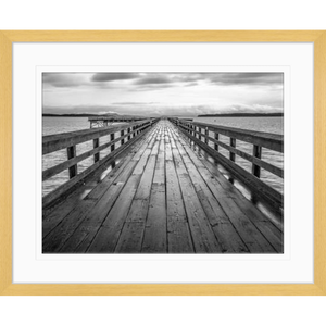 Black and white wall art features a long pier cascading into the deep ocean, with an array of white clouds overhead in an oak frame