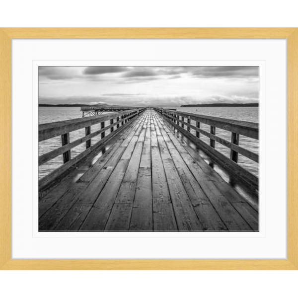 Black and white wall art features a long pier cascading into the deep ocean, with an array of white clouds overhead in an oak frame