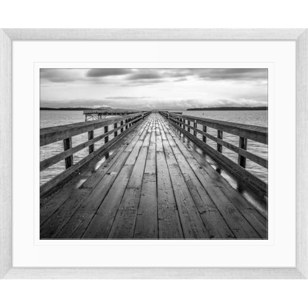 Black and white wall art features a long pier cascading into the deep ocean, with an array of white clouds overhead in a silver frame