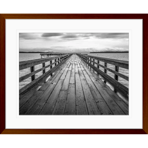 Black and white wall art features a long pier cascading into the deep ocean, with an array of white clouds overhead in a wood grain frame