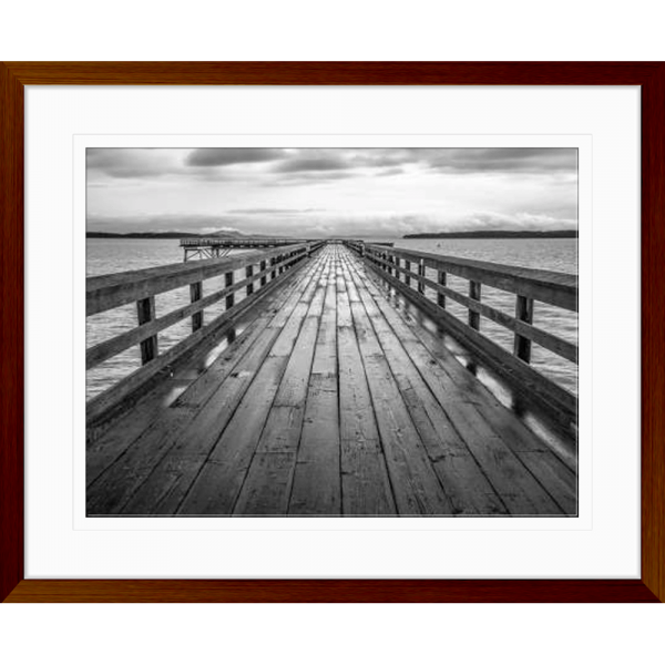 Black and white wall art features a long pier cascading into the deep ocean, with an array of white clouds overhead in a wood grain frame