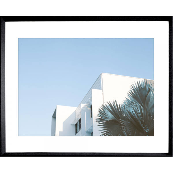 Black framed Tropical Wall Art - The Weekender - Artist Collection, featuring striking a crisp white tropical hotel nestled amongst breezy palm trees, against a sky blue background. 