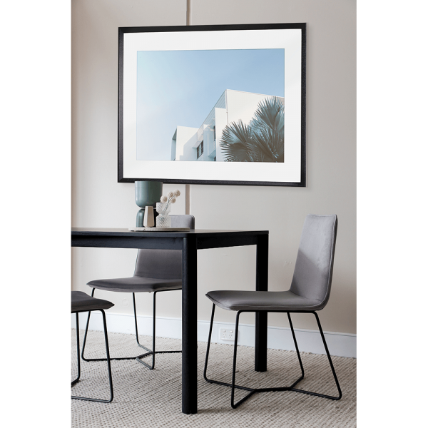 Black framed mounted in the lounge - Tropical Wall Art - The Weekender - Artist Collection, featuring striking a crisp white tropical hotel nestled amongst breezy palm trees, against a sky blue background. 