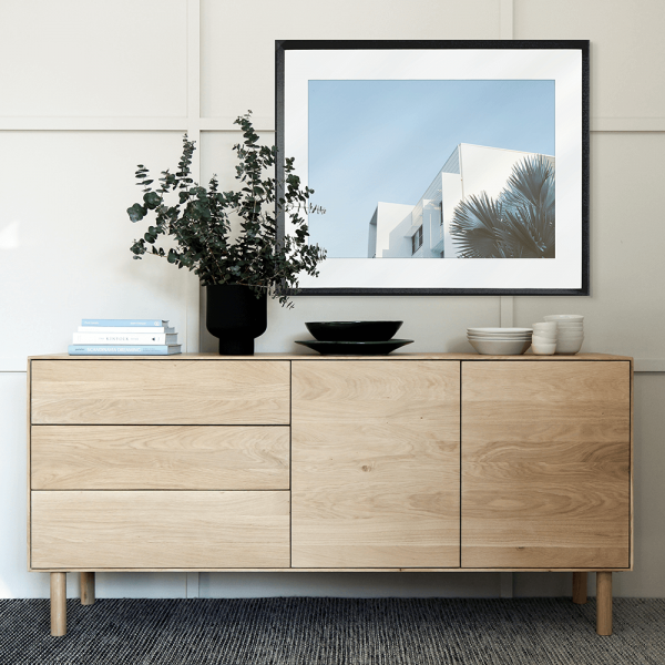 Black framed mounted Tropical Wall Art - The Weekender - Artist Collection, featuring striking a crisp white tropical hotel nestled amongst breezy palm trees, against a sky blue background. 