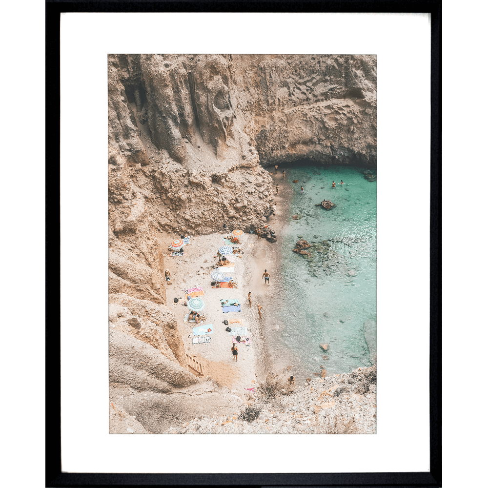 Black framed Coastal Wall Art - Vacanza 01 – Studio Collection, Featuring a relaxing beach scene nestled within a cliff face. Clear aqua water with beach lovers sun baking in the sunshine.