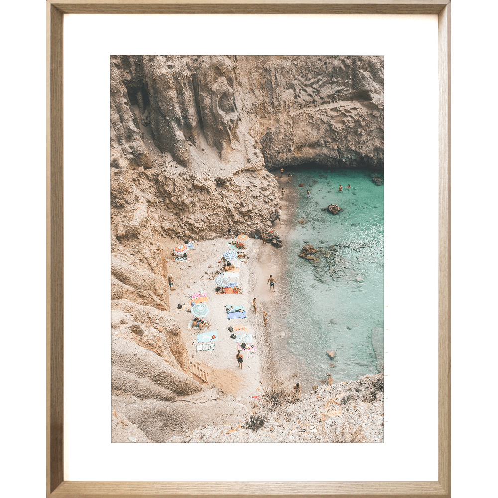 Gold framed Coastal Wall Art - Vacanza 01 – Studio Collection, Featuring a relaxing beach scene nestled within a cliff face. Clear aqua water with beach lovers sun baking in the sunshine.