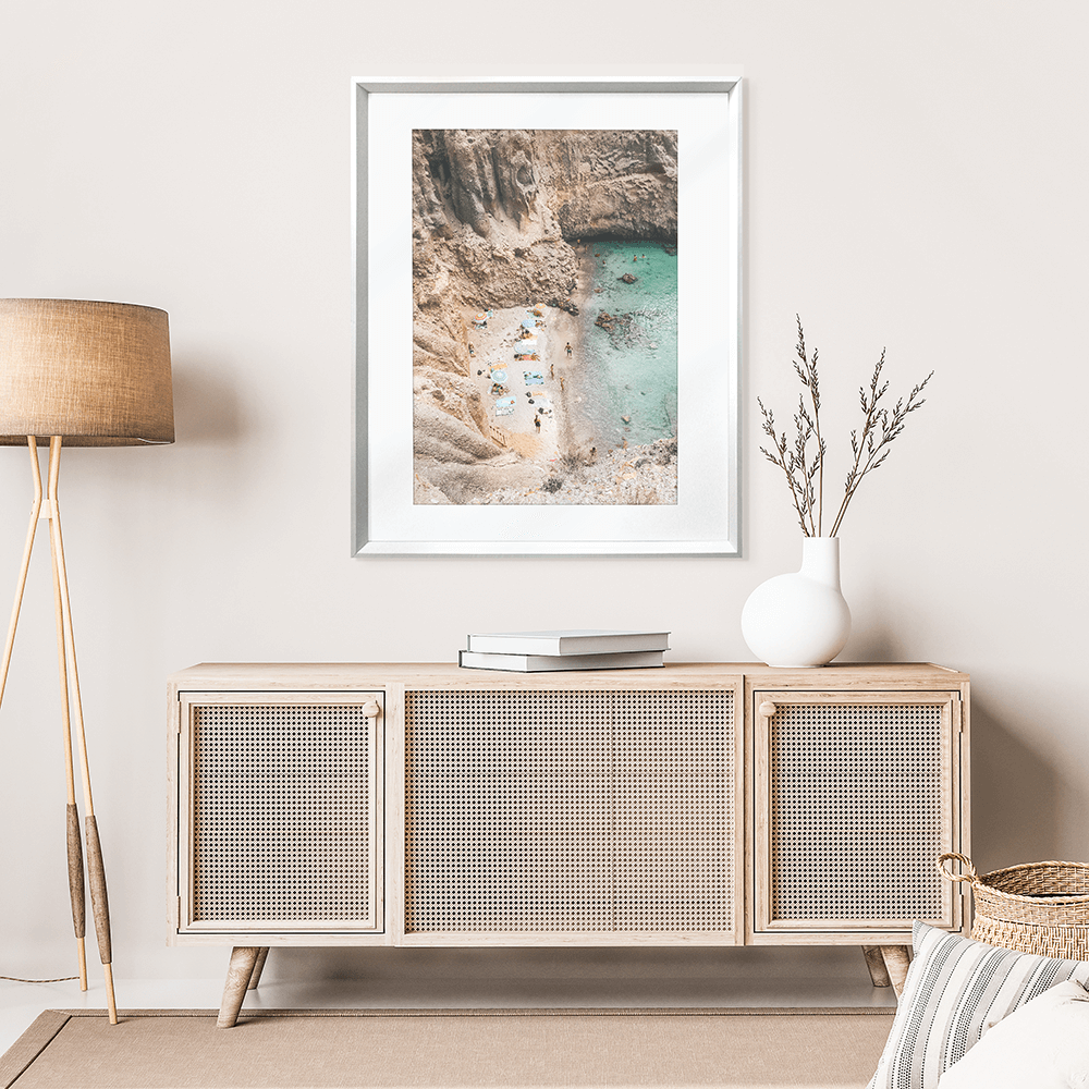 Wall mounted Silver framed Coastal Wall Art - Vacanza 01 – Studio Collection, Featuring a relaxing beach scene nestled within a cliff face. Clear aqua water with beach lovers sun baking in the sunshine.