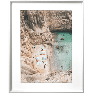 Silver framed Coastal Wall Art - Vacanza 01 – Studio Collection, Featuring a relaxing beach scene nestled within a cliff face. Clear aqua water with beach lovers sun baking in the sunshine.